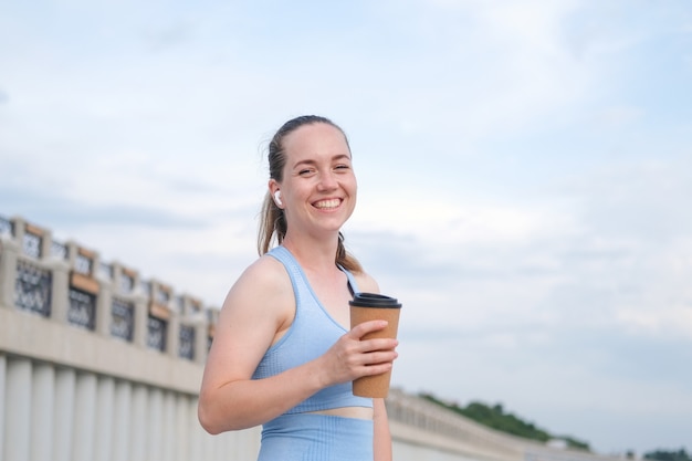 Młoda szczęśliwa kobieta fitness działa w mieście podczas słuchania muzyki w słuchawkach, trzymając filiżankę kawy. Zdjęcie wysokiej jakości