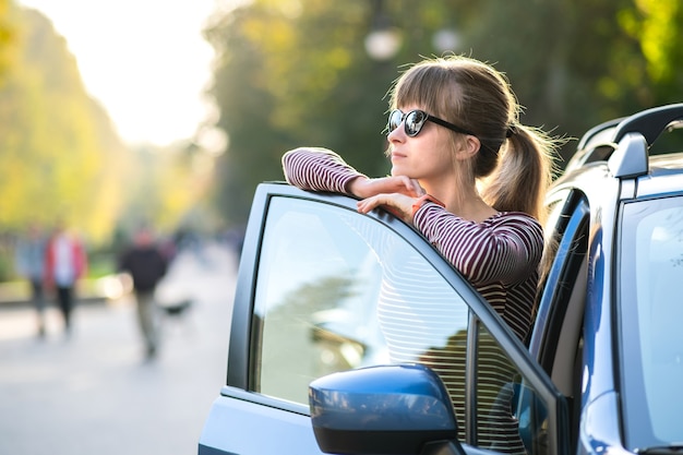 Młoda szczęśliwa kobieta kierowca stojący w pobliżu jej samochodu na ulicy miasta w lecie. Cele podróży i koncepcja transportu.