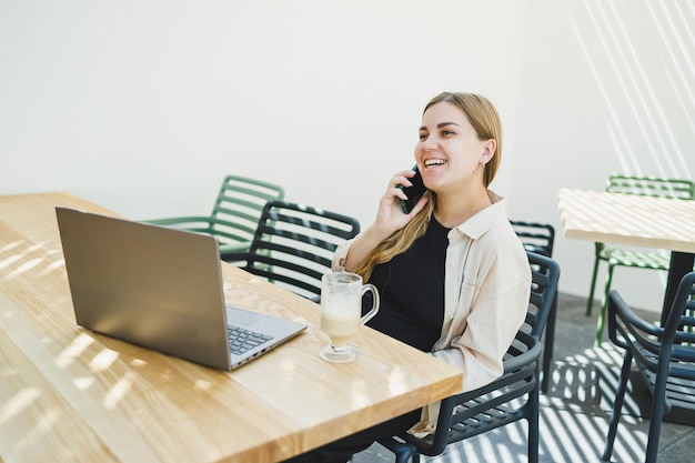 Młoda szczęśliwa kobieta siedzi przy stoliku kawiarni na świeżym powietrzu i rozmawia przez telefon z filiżanką kawy uśmiechnięta kobieta cieszy się pracą zdalną w kawiarni lub studiuje online na laptopie