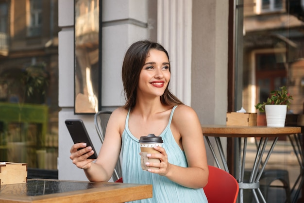 młoda szczęśliwa kobieta siedzi w kawiarni na świeżym powietrzu podczas korzystania z telefonu komórkowego i picia kawy
