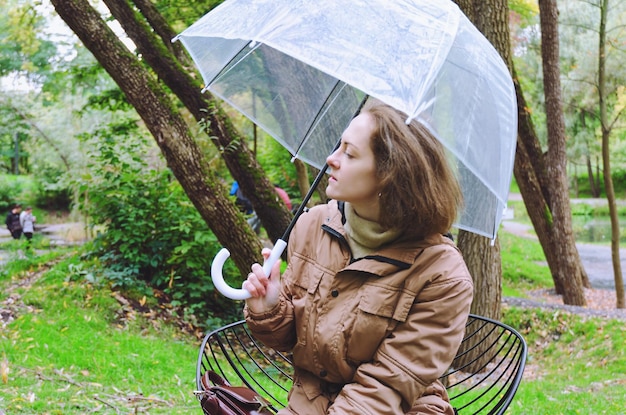 Młoda szczęśliwa kobieta spaceru w deszczu w zielonym parku z parasolem, uśmiechnięta, zabawa. osoba siedzi na ławce