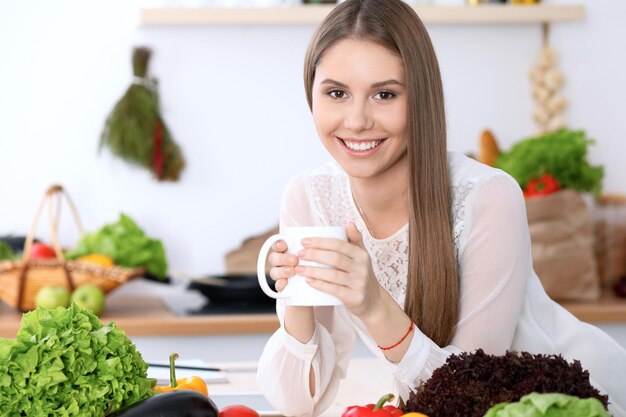 Młoda szczęśliwa kobieta trzyma białą filiżankę i patrzy w kamerę siedząc przy drewnianym stole w kuchni wśród zielonych warzyw
