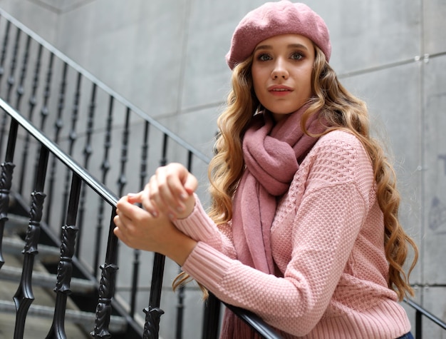 Młoda szczęśliwa kobieta z długimi kręconymi włosami, ubrana w różowy szalik i kapelusz na co dzień stoi przy schodach