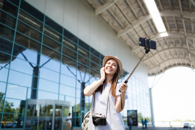 Młoda szczęśliwa ładna podróżniczka turystyczna kobieta z retro aparatem fotograficznym robi selfie na telefonie komórkowym z samolubnym kijem na lotnisku