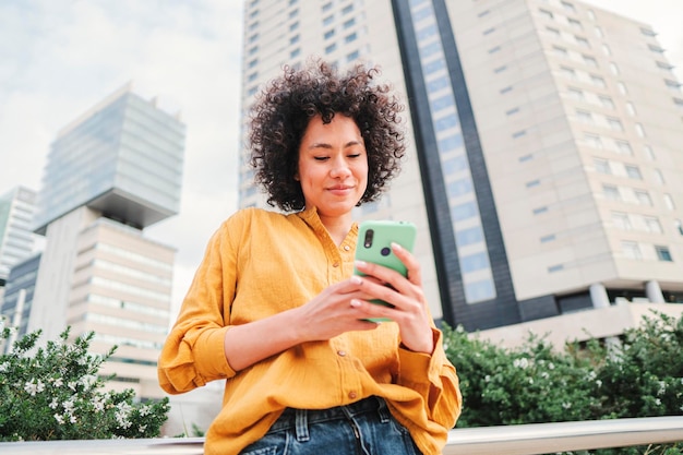 Młoda szczęśliwa latynoska kobieta ogląda śmieszne filmy w aplikacji społecznościowej smartfona na zewnątrz Latynoska dama w żółtej koszuli wysyłająca SMS-y z telefonem komórkowym na ulicy
