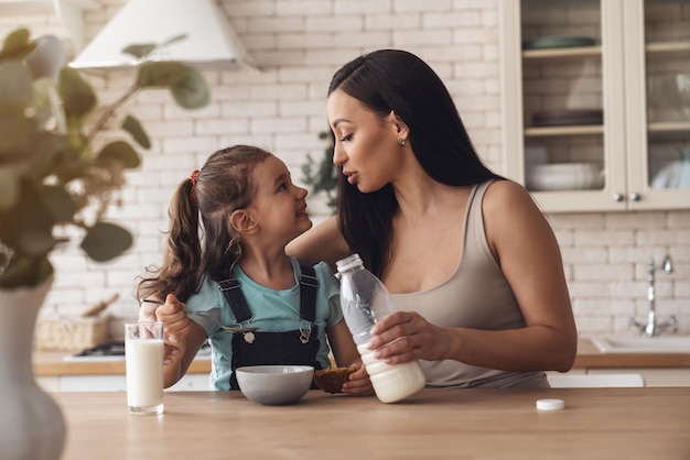 Młoda szczęśliwa mama przygotowuje śniadanie dla swojej córeczki, dodając mleko i musli do talerza, siedząc przy stole w kuchni w domu