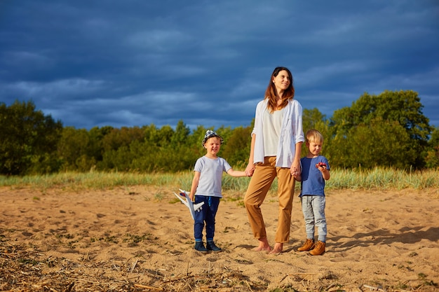 Zdjęcie młoda szczęśliwa matka z dwoma chłopcami bawiącymi się na piasku w pobliżu lasu, w rękach jednego z nich model samolotu cywilnego i kapelusz na głowie