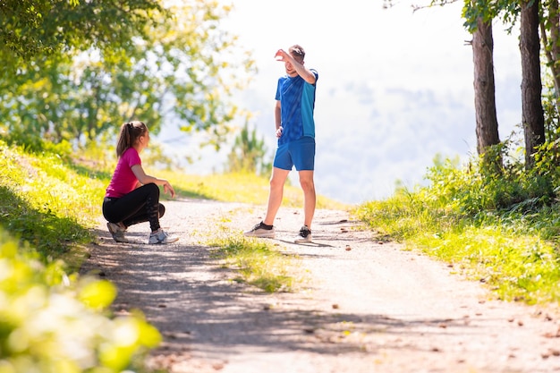 młoda szczęśliwa para ciesząca się zdrowym stylem życia, rozgrzewająca się i rozciągająca przed joggingiem na wiejskiej drodze przez piękny słoneczny las, koncepcja ćwiczeń i fitness