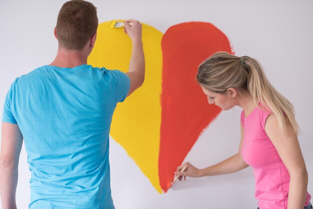 Młoda szczęśliwa para maluje serce na ścianie podczas naprawy w domu.