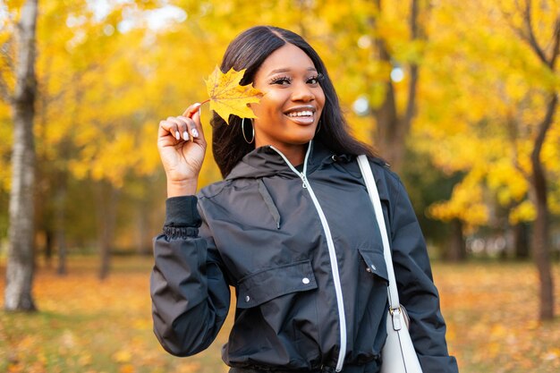 Młoda szczęśliwa piękna czarna dziewczyna ze słodkim uśmiechem w modnych ubraniach na co dzień trzymająca żółty jesienny liść spaceruje po parku z jasnymi kolorowymi jesiennymi liśćmi