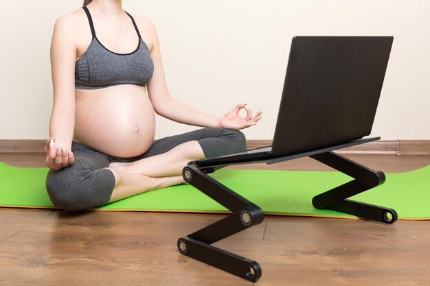 Młoda szczęśliwa zdrowa joga fitness piękna kobieta w ciąży robi trening jogi bakasana na macie do jogi naprzeciwko laptopa z koncepcją stylu życia online klasy mistrzowskiej w czasie koronawirusa