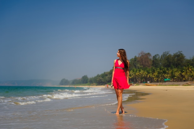 Młoda szczupła długowłosa brunetka w czerwonej sukience chodzi boso po tropikalnej plaży wzdłuż fal na tle morza i niebieskiego nieba w upalny dzień. W oddali zielone wzgórze i ludzie na brzegu.