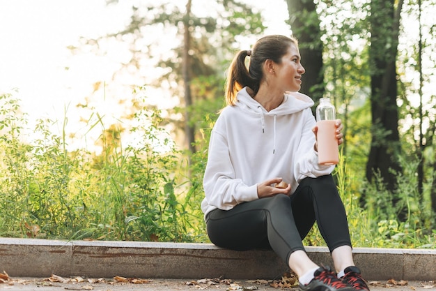 Młoda szczupła kobieta brunetka w sportowych ubraniach biega i woda pitna w jesiennym parku w złotej godzinie czas wschodu słońca Zdrowie i dobre samopoczucie fitness styl życia