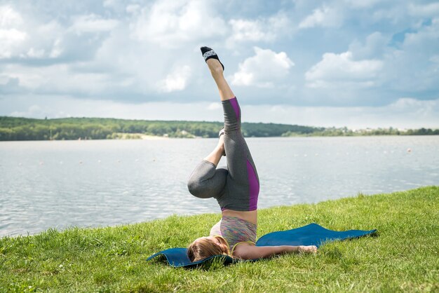 Młoda szczupła kobieta nosi ubrania fitness, robi ćwiczenia gimnastyczne na macie do jogi w pobliżu jeziora. Pojęcie zdrowego stylu życia