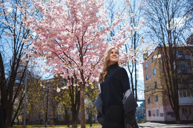 Młoda, szczupła modelka z długimi falującymi włosami i ubrana w szary płaszcz, trampki, stoi na ulicy w pobliżu kwitnącego krzewu z pięknymi różowymi kwiatami w tle i pozami.