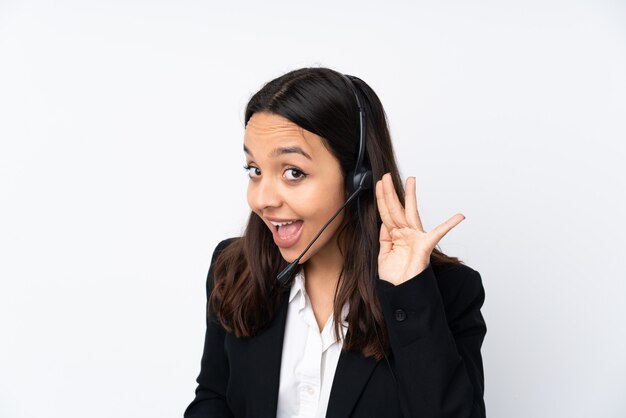 Młoda telemarketer kobieta odizolowywająca na biel ścianie słucha coś kładąc rękę na ucho
