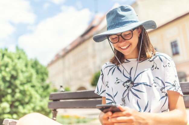 Młoda turystka uśmiechająca się do swojego telefonu komórkowego, siedząca na ławce w centrum starego miasta.