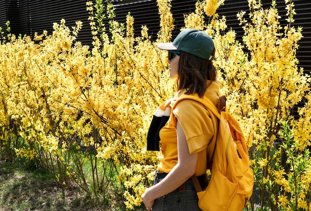 Młoda turystka w czapce z żółtym plecakiem wśród kwitnących krzewów forsycji wiosną lub latem