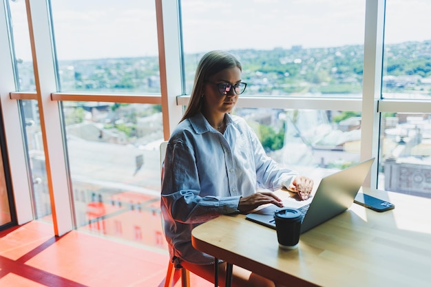 Młoda tysiącletnia kobieta w klasycznych okularach ochronnych łączy się z internetem WIFI na laptopie w przestrzeni coworkingowej, aby przeglądać informacje internetowe na cyfrowych netbookach europejska kobieta na czacie