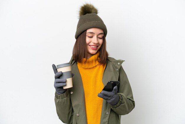 Młoda Ukrainka ubrana w zimową kurtkę na białym tle trzymająca kawę na wynos i telefon komórkowy