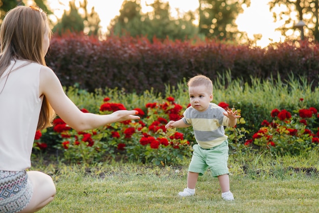 Młoda Urocza Mama Pomaga I Uczy Swojego Małego Synka Stawiać Pierwsze Kroki Podczas Zachodu Słońca W Parku Na Trawie.