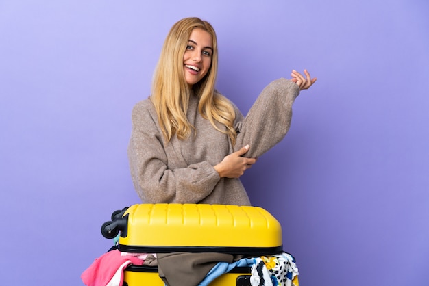 Młoda urugwajska blondynka z walizką pełną ubrań na izolowanej fioletowej ścianie wyciągającej ręce na bok, zapraszając do siebie