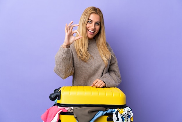 Młoda Urugwajska blondynka z walizką pełną ubrań na odosobnionej fioletowej ścianie pokazuje znak ok palcami
