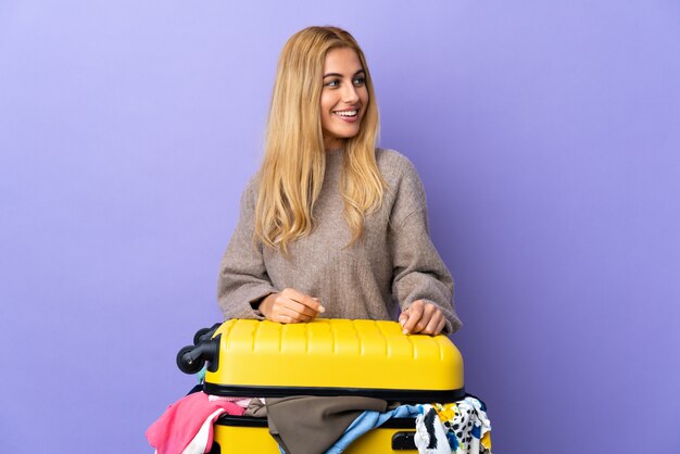 Młoda Urugwajska blondynki kobieta z walizką pełną ubrania nad odosobnioną purpurą izoluje patrzeć z boku i ono uśmiecha się