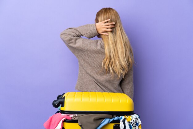 Młoda urugwajska blondynki kobieta z walizką pełną ubrania nad purpurową ścianą w tylnym położeniu i myśleniu