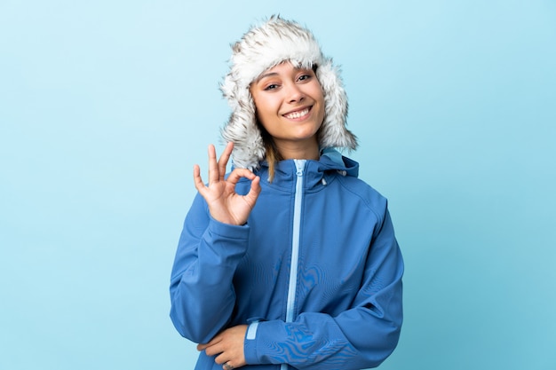 Młoda Urugwajska dziewczyna z zima kapeluszem odizolowywającym na błękitnym tle pokazuje ok znaka z palcami
