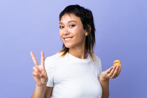 Młoda Urugwajska Kobieta Na Odosobnionych Purpurach Trzyma Kolorowych Francuskich Macarons I Pokazuje Zwycięstwo Znaka