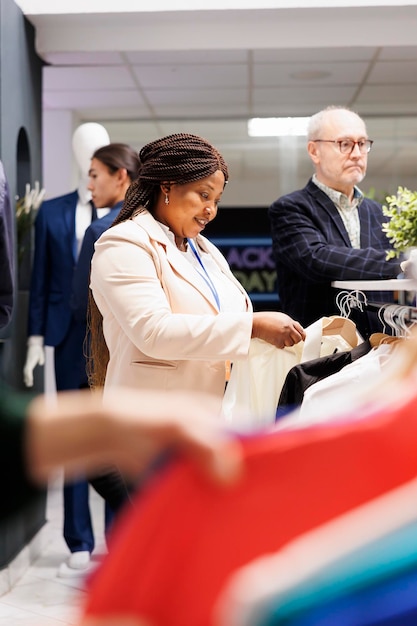 Młoda uśmiechnięta afroamerykańska asystentka sprzedaży stojąca w sklepie z odzieżą układająca ubrania na półce, ciesząca się pracą w przemyśle detalicznym, kierownik merchandising pracujący w centrum handlowym