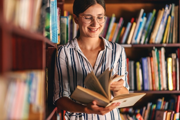 Młoda uśmiechnięta atrakcyjna dziewczyna college'u, opierając się na półkach z książkami w bibliotece i szukając materiału do pracy domowej.