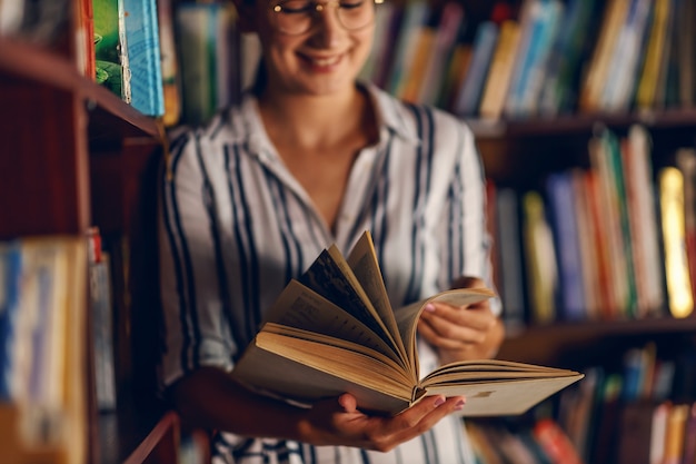 Młoda uśmiechnięta atrakcyjna dziewczyna college'u, opierając się na półkach z książkami w bibliotece i szukając materiału do pracy domowej.