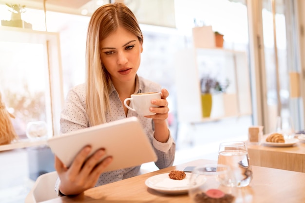 Młoda uśmiechnięta bizneswoman na przerwie w kawiarni. Pije kawę i używa tabletu.