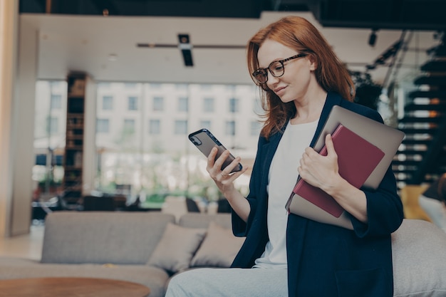 Młoda uśmiechnięta bizneswoman w okularach z laptopem i agendą pod pachą, patrząc na smartfona