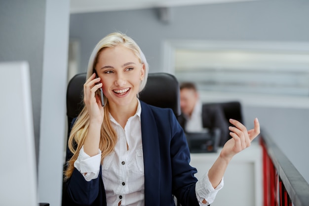 Młoda uśmiechnięta blondynka kaukaski kobieta siedzi w biurze i za pomocą inteligentnego telefonu do rozmowy biznesowej. Bez walki nie ma postępu.