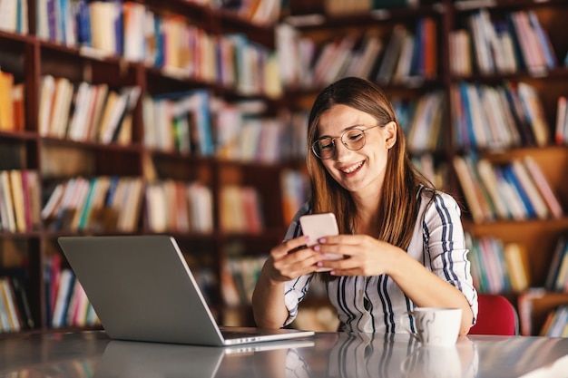 Młoda Uśmiechnięta Brunetka Z Okularami Siedzi W Bibliotece I Za Pomocą Inteligentnego Telefonu. Na Stole Jest Laptop. Koncepcja Zdalnego Uczenia Się.