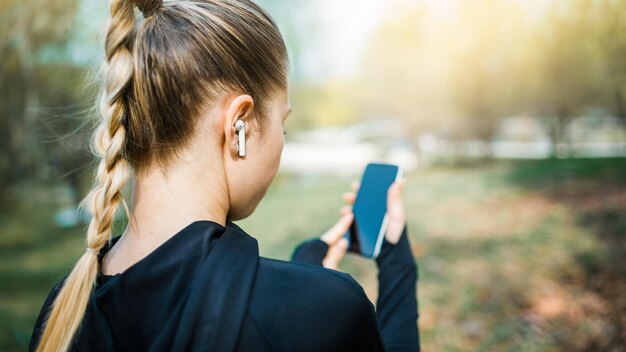 Młoda uśmiechnięta dziewczyna uprawiająca sport i biegająca w parku używając telefonu do słuchania muzyki z bezprzewodowymi słuchawkami na zachodzie słońca w mieście oglądając ekran