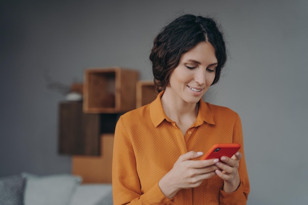 Młoda Uśmiechnięta Hiszpańska Dama Trzyma Smartfon Patrząc Na Ekran Z Uśmiechem I Wysyłając Sms
