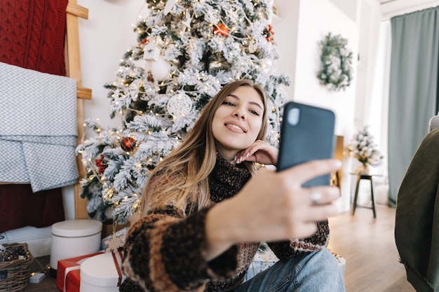 Młoda uśmiechnięta kaukaski kobieta robi selfie przy użyciu telefonu komórkowego w pobliżu choinki w domu.