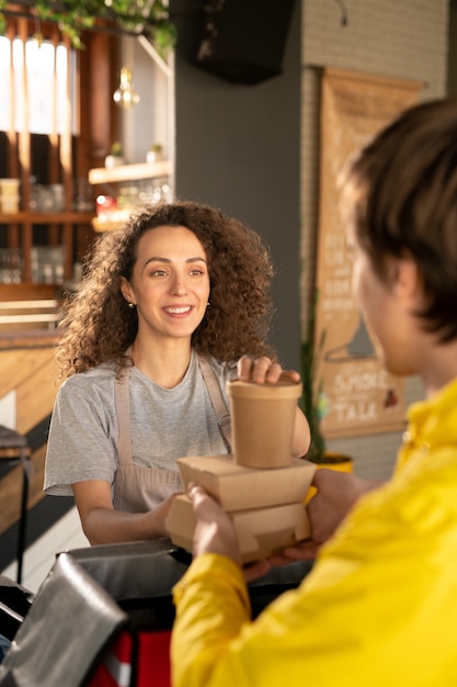 Młoda uśmiechnięta kelnerka patrząc na kuriera stojącego przed nią, pomagając mu pakować zamówienia klientów do dużej torby w restauracji