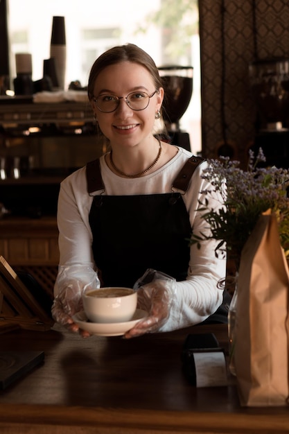 Młoda uśmiechnięta kobieta barista rozdaje filiżankę cappuccino klientowi w kawiarni