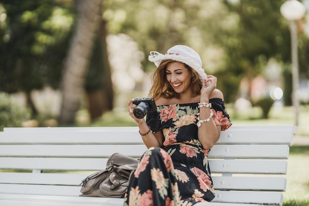 Młoda uśmiechnięta kobieta, patrząc na zdjęcia aparatem cyfrowym i ciesząc się siedząc na ławce w parku.