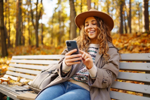 Młoda uśmiechnięta kobieta siedząca na ławce w parku jesieni używająca telefonu w stylowych ubraniach w szczęśliwym nastroju