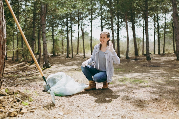 Młoda uśmiechnięta kobieta sprzątanie śmieci do worków na śmieci Wyświetlono w porządku znak w parku lub lesie. Problem zanieczyszczenia środowiska