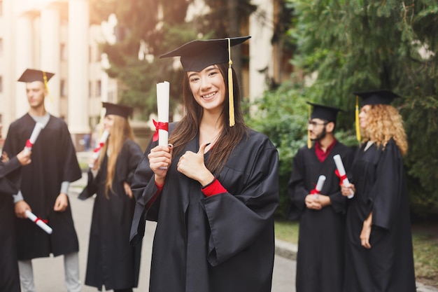 Młoda uśmiechnięta kobieta w dniu ukończenia studiów pokazując kciuk do góry stojący z wieloetniczną grupą studentów. Koncepcja edukacji, kwalifikacji i sukni.