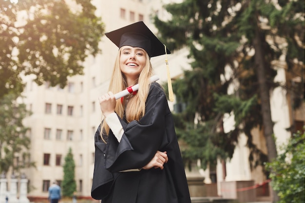 Młoda uśmiechnięta kobieta w dniu ukończenia studiów w uniwersytecie posiadającym dyplom. Koncepcja edukacji, kwalifikacji i sukni.