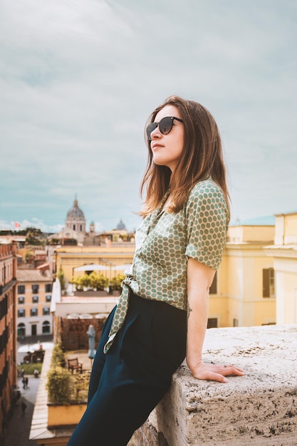 Zdjęcie młoda uśmiechnięta kobieta w okularach przeciwsłonecznych siedzi na dachu z panoramicznym widokiem na rzym we włoszech