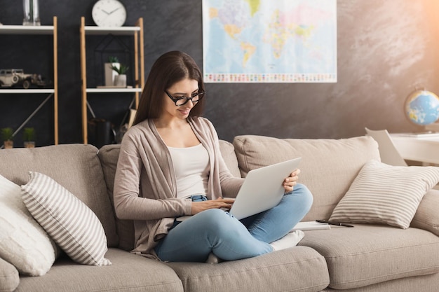 Młoda uśmiechnięta kobieta w okularach za pomocą laptopa w domu na kanapie. Wesoła dziewczyna pisząca na komputerze, edukacji lub freelance, kopia przestrzeń